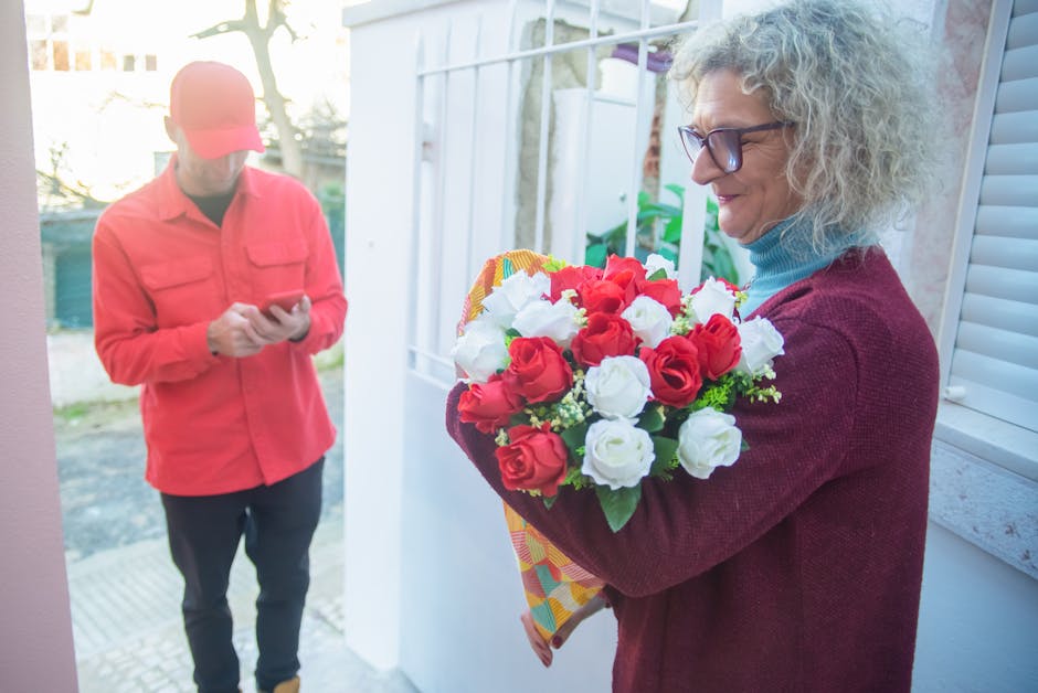 A woman with curly gray hair and glasses, wearing a maroon sweater and blue turtleneck, is holding a floral bouquet featuring red and white roses, wrapped in decorative paper. She stands near a white 