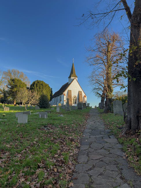 The image depicts a small white chapel with a pointed steeple set atop a grassy hill, surrounded by a few leafless trees and scattered gravestones in a cemetery. A narrow, uneven stone path leads up t
