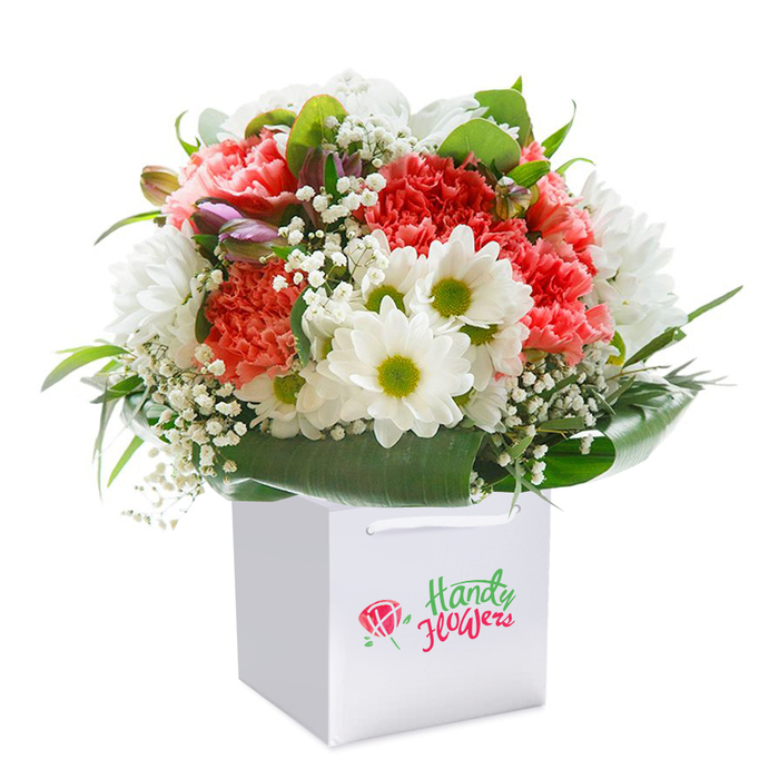 Presented upright in a crisp white square gift bag, this luminous bouquet captures the vibrant character of a West Drayton celebration. Rich coral, peach, and vivid scarlet carnations-blooming with layers of exuberant, ruffled petals-immediately evoke feelings of warmth and affection. Nestled among them, bright white, daisy-like blooms shine, their chartreuse centers providing a lively counterpoint and recalling sunny afternoons at the local park. Wispy white sprigs of baby's breath are scattered throughout the design, offering a cloud-like softness that brings a sense of space and airiness. A subtle cluster of purple-tinted, closed buds is tucked on one side, contributing an element of intrigue and a subtle pop of colour. Encircling these blossoms, strong, deep green leaves arc outward to cradle the arrangement, delivering structure and classic form. The gift bag is both elegant and practical, with smooth ribbon handles perfectly suited for floral delivery along West Drayton's winding streets. While a colourful emblem sits at the fore, the focus remains entirely on the freshness and skillful composition of the blooms themselves. Every detail is heightened by the white background, suggesting purity and a standard of quality that sets West Drayton's florists apart. It's the sort of bouquet suited for a milestone, a heartfelt thank you, or brightening someone's home along the banks of the Grand Union Canal.